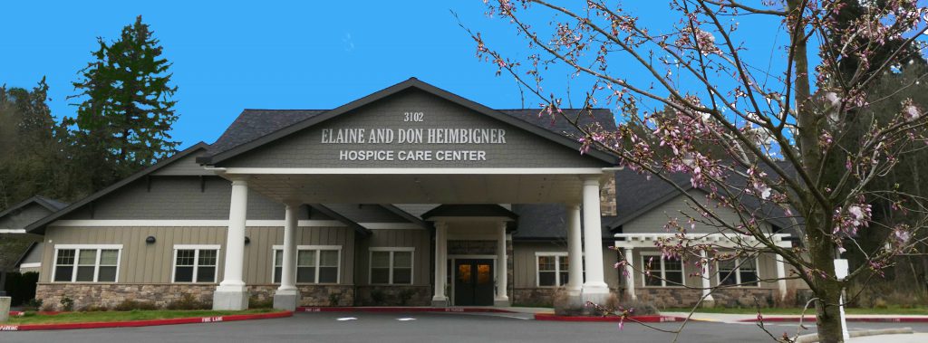 Front entrance of the Elaine and Don Heimbigner Hospice Care Center with a blue sky and a blooming dogwood tree